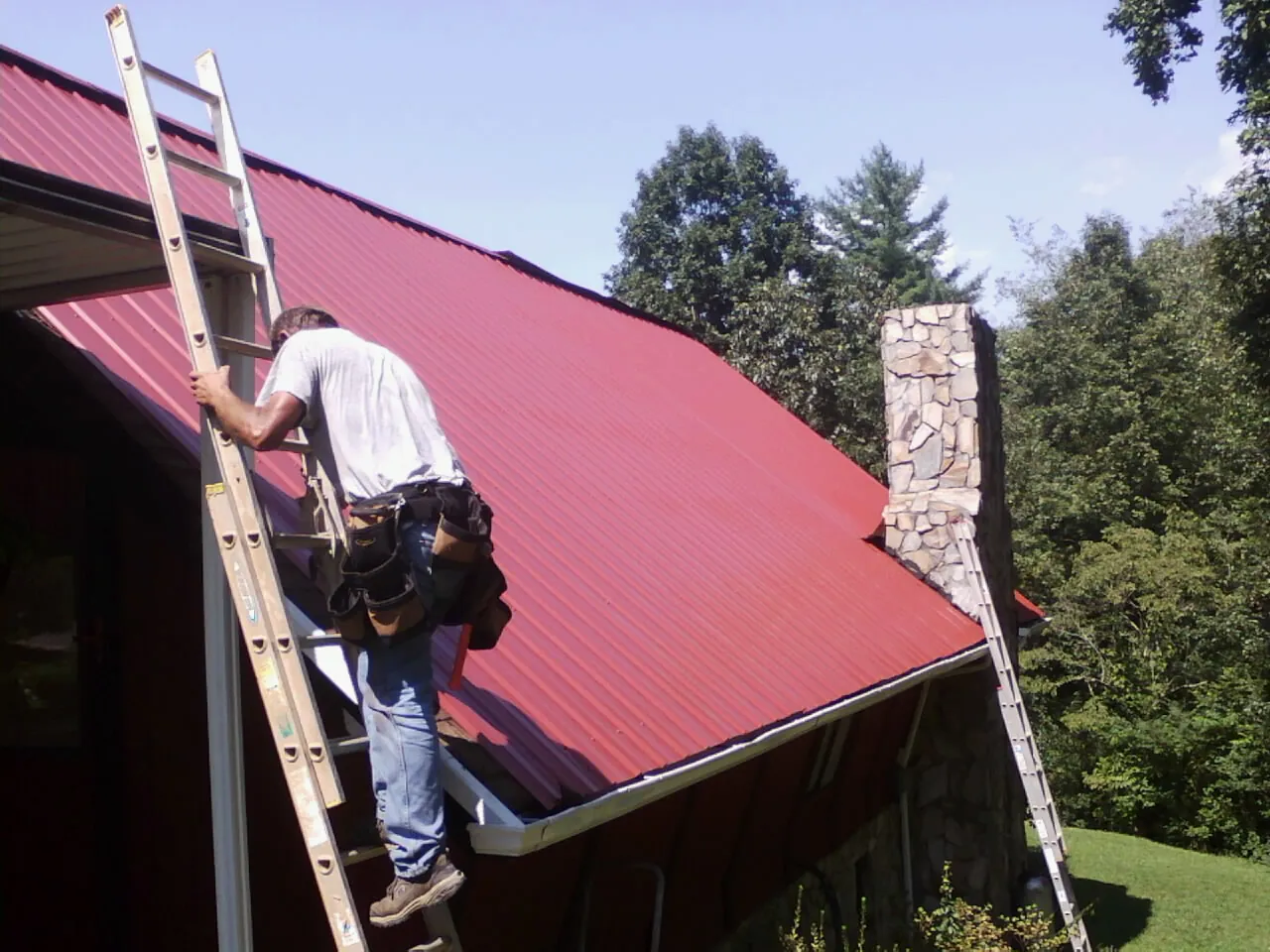 Roofer on a ladder beside a red metal roof and stone chimney — Soffit & Fascia Repair by Roofing Brothers in Minerva